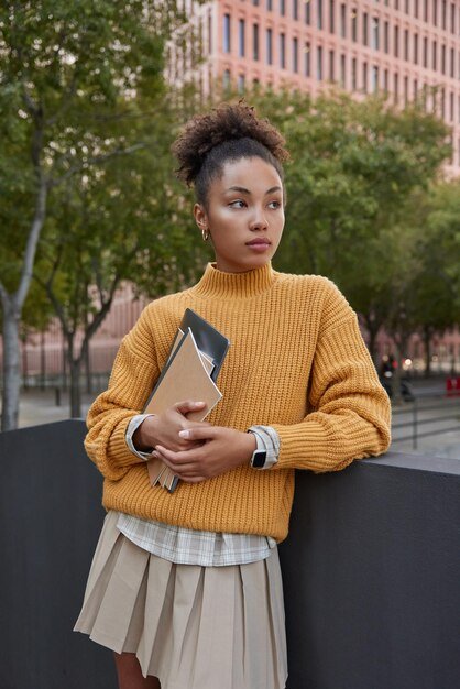 vertical-shot-thoughtful-curly-female-student-holds-digital-tablet-notebook-goes-university-wears-knitted-yellow-sweater-skirt-focused-away-stands-outdoors-against-urban-background_273609-60487.jpg