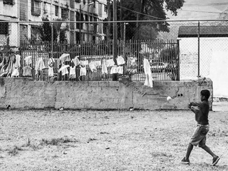 foto blanco y negro de un niño  no mayor de 10 años  en un terreno baldío, vestido con franela y pantalones cortos sujetando un bate de beisbol en la mano mirando una pelota en el aire. Alrededor bloques de viviendas, un estacionamiento y ropa colgando de las ventanas