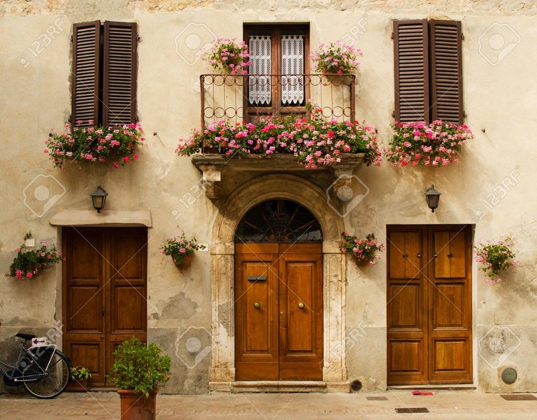 92517047-fachada-de-la-casa-antigua-en-pienza-con-ventanas-balcón-con-flores-puertas-de-madera-plantas-toscana-i.jpg