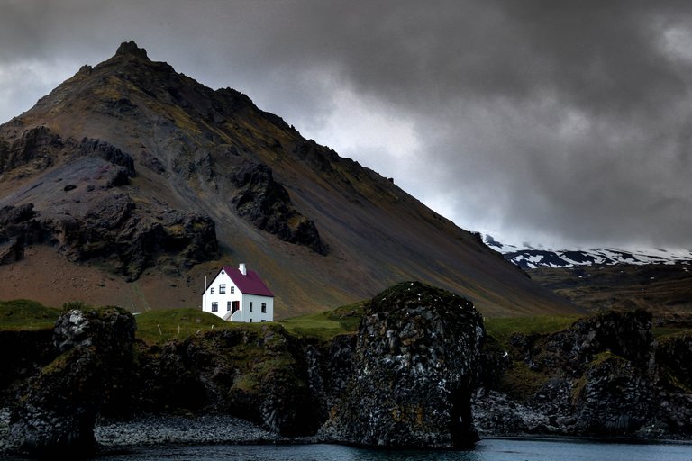 A sturdy white house on a rocky outcrop along the sea beside a mountain. In the background a storm is moving in.