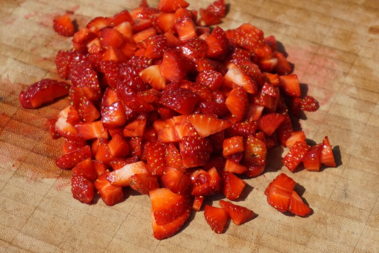 Wooden chopping board on which small cut strawberries are piled on top.