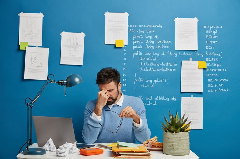 exhausted-businessman-rubs-nose-takes-off-spectacles-suffers-from-eye-strain-headache-has-problems-work-sits-coworking-space-with-laptop-computer-blue-wall-with-written-notes.jpg