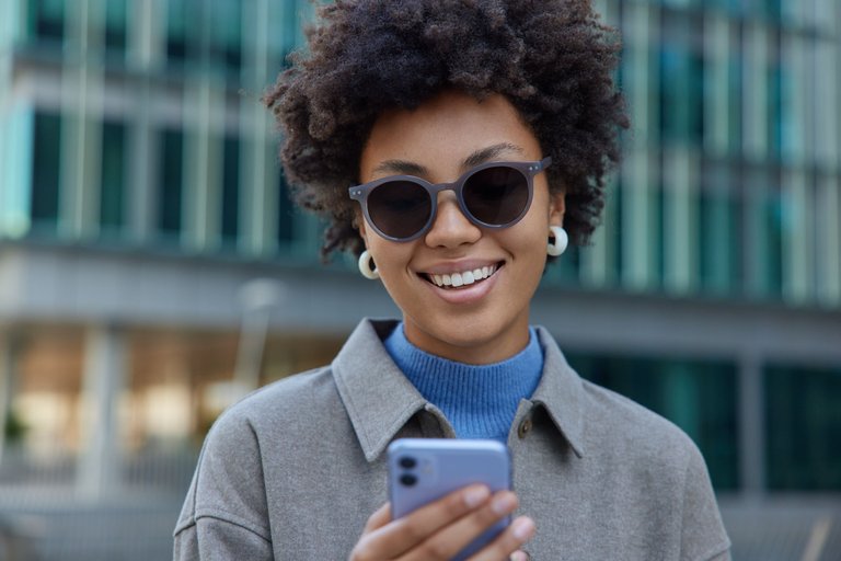 smiling-lady-with-afro-hair-bowses-mobile-phone-while-standing-city-street-being-good-mood-wears-sunglasses-coat-surfs-social-networks-connected-wireless-internet.jpg