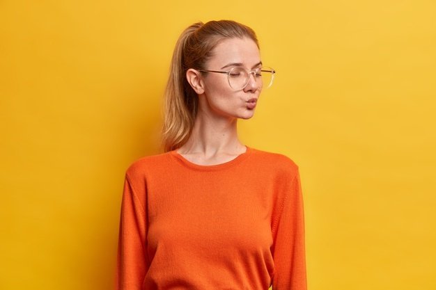 beautiful-young-european-woman-stands-with-closed-eyes-keeps-lips-rounded-has-romantic-mood-pony-tail-wears-orange-jumper_273609-43593.jpg