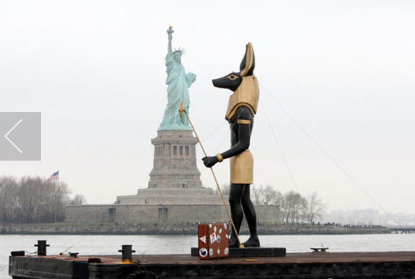 25foot tall replica statue of the Egyptian god Anubis, with a suitcase at his feet, passes in front of the Statue of Liberty 323 233 2010.png