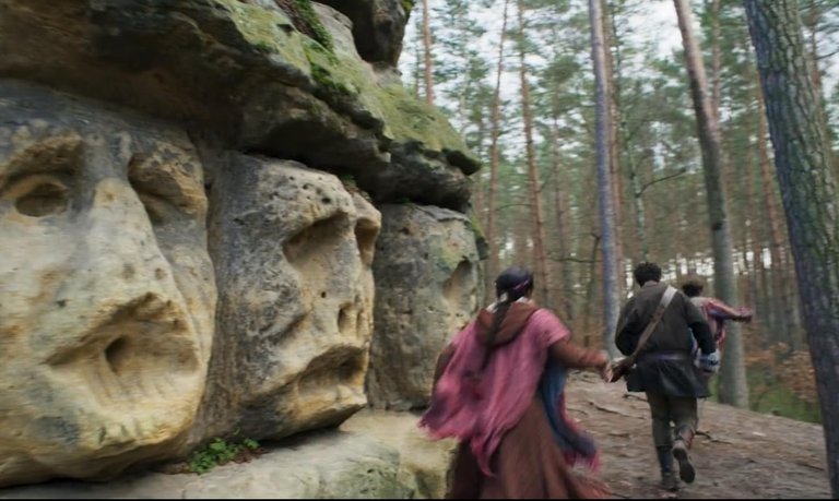 Screenshot of three people running near a worn-out ancient monument carven into the stone of the cliff face