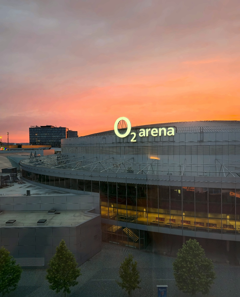 O2 Arena with its illuminated sign and curved roof under a pink-orange sunset sky in Prague.