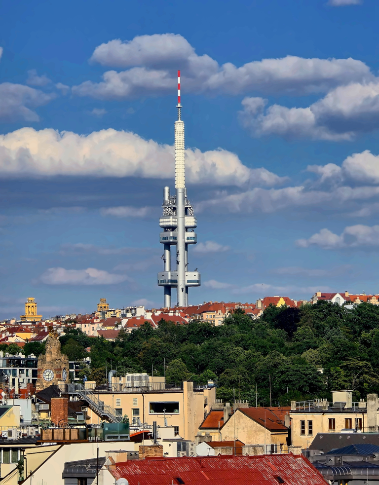 Žižkov Television Tower rises above Prague’s red-roofed buildings and leafy trees under a blue sky.