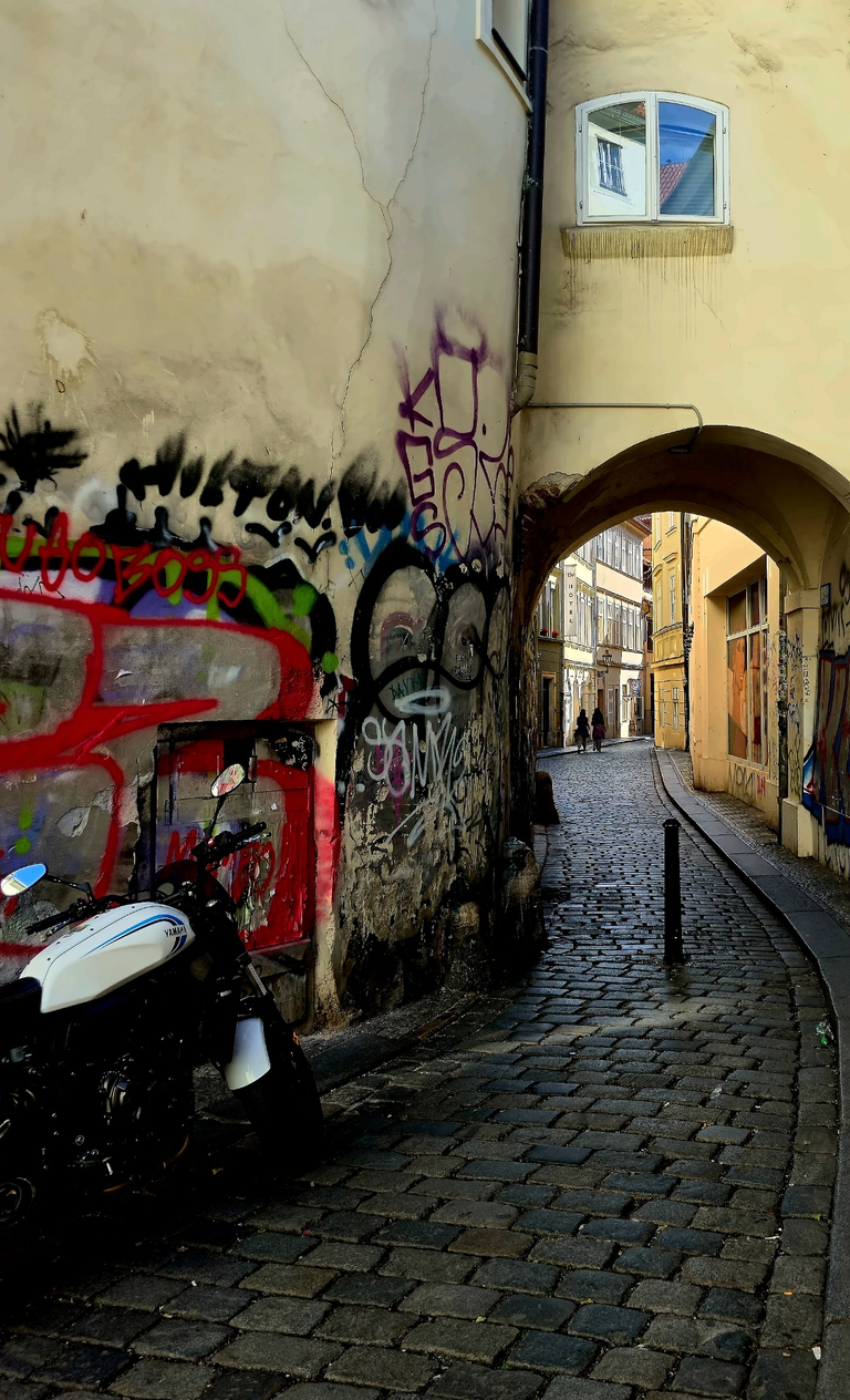 Graffiti-covered passage with a parked scooter and cobblestones beneath an arch in Prague's Staré Město.