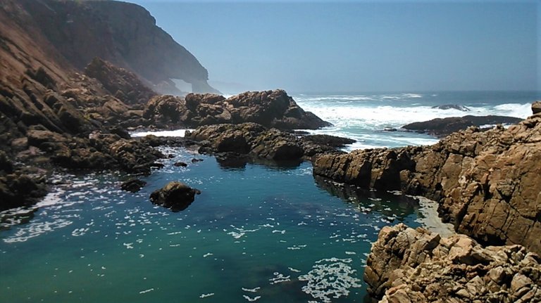 A calm pool fed by the rougher sea just other side of the rocky boundary provided by nature with epic arch in the distance.