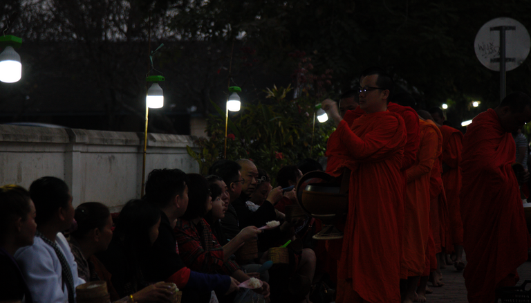 People sit along the road while giving food for the monk that pass by