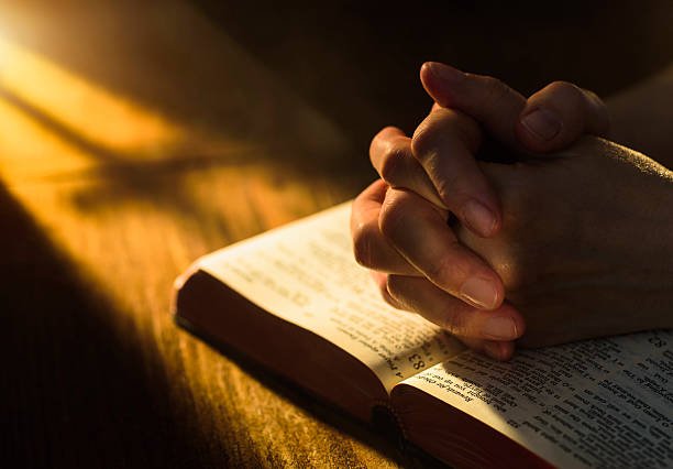 close-up-of-hands-clasped-on-open-bible-picture-id562612565.jpeg