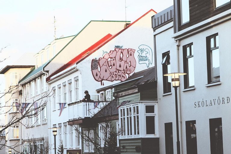 An elderly woman standing on her balcony above a tourist-filled street. She’s standing in front of an "Ugly Boys" graffiti tag.