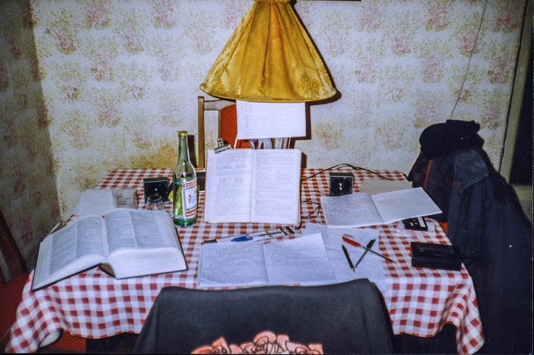 A kitchen table with a dictionary and some notebooks and pens and a bottle of vodka`