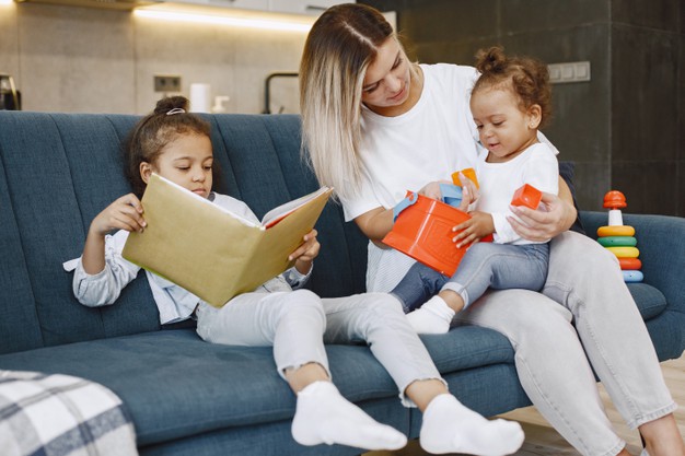 madre-e-hijos-relajandose-juntos-sofa-casa-sala-estar-ninas-leyendo-libro-jugando-juguetes_1157-48360.jpg