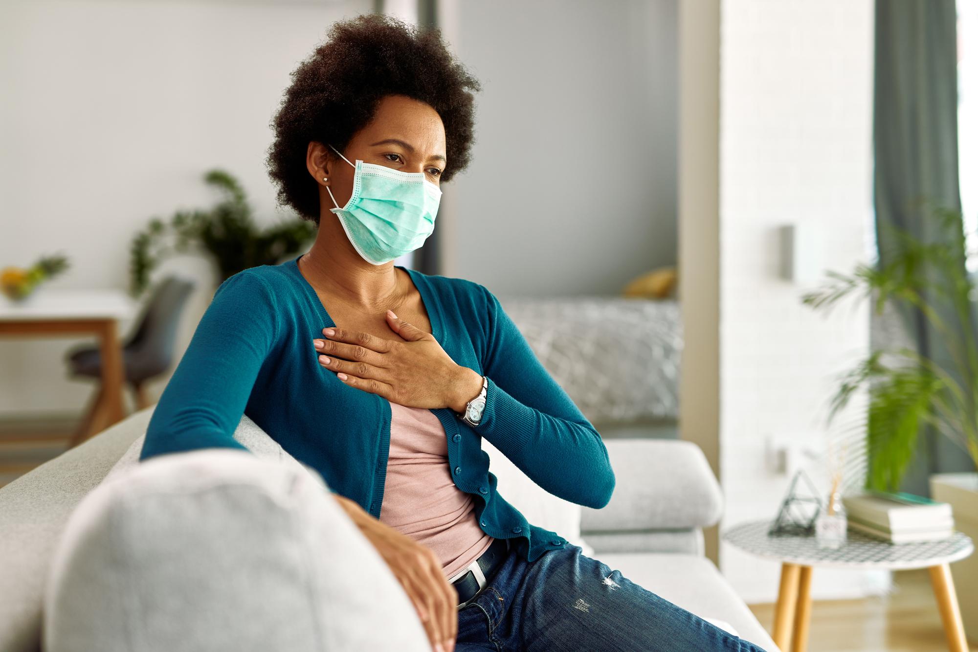 black-woman-wearing-face-mask-holding-her-chest-pain-while-sitting-sofa-living-room.jpg